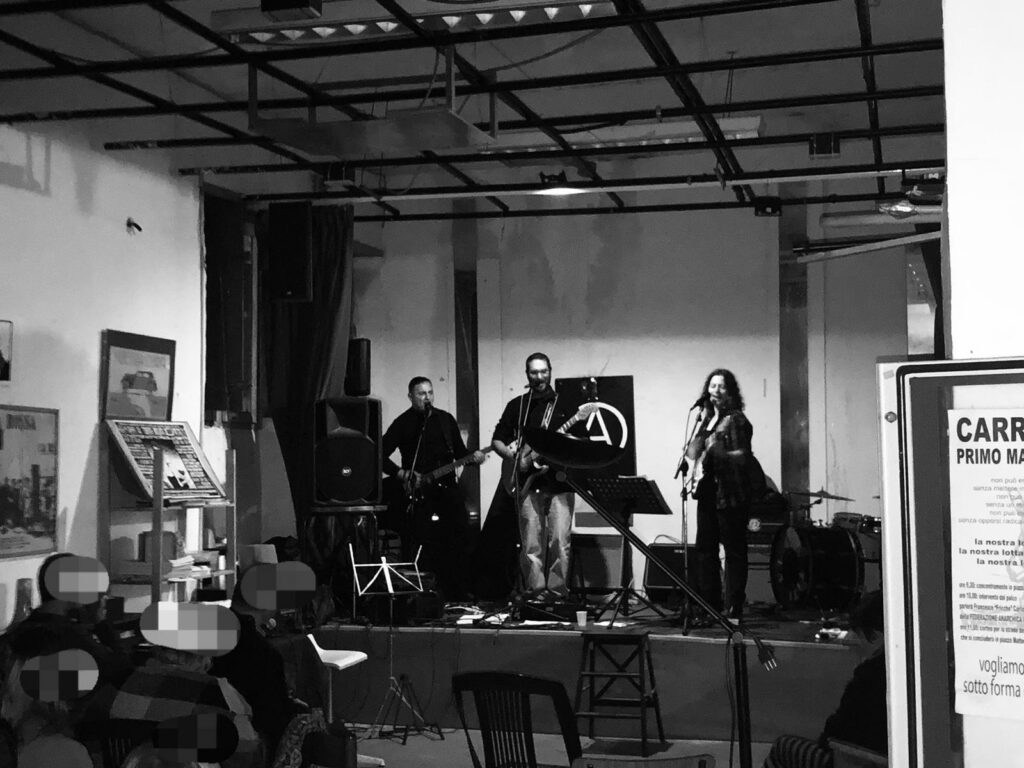 foto di bassista, chitarrista e secondo chitarrista sul palco. c'è gente del pubblico col viso pixelato