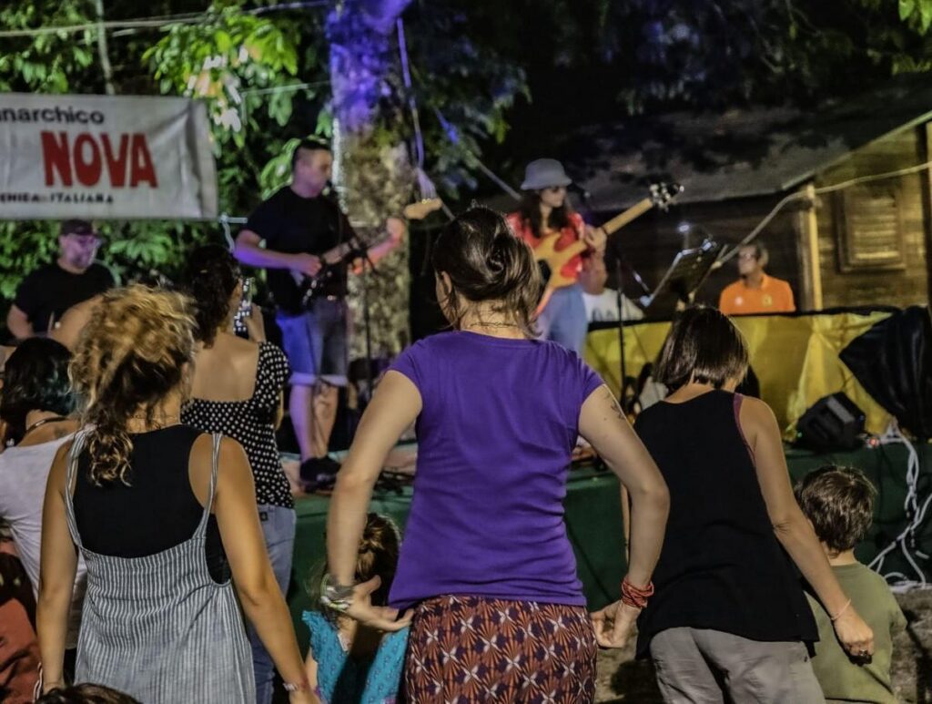 chitarrista, bassista e batterista sul palco. in primo piano alcune persone del pubblico prese di schiena. è sera, in alto si intravede lo striscione di umanità nova